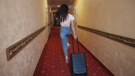 happy female traveler with suitcase indoors. young woman in casual clothes walking along the hotel corridor with trolley bag and jumping joyfully. vacation concept.