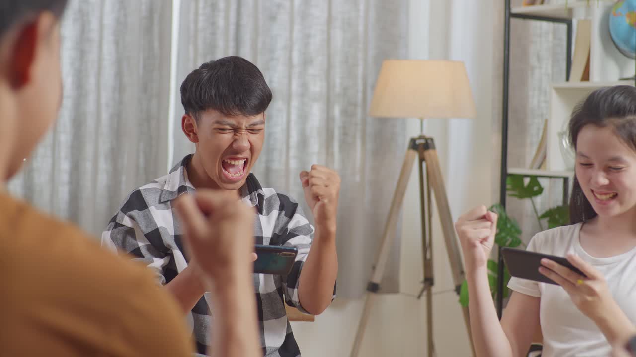 아시아 십대 그룹이 의자에 앉아서 집에서 스마트폰으로 게임을 하는 원을 형성합니다. 성공을 축하하고 게임 경기에서 승리하고, 하이 파이브 제스처를 합니다.