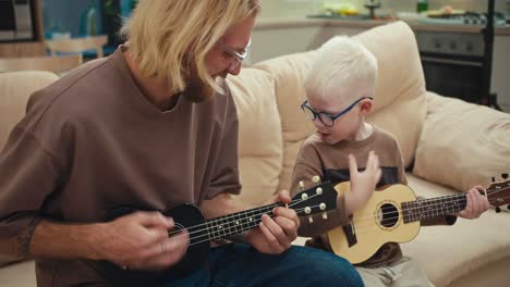 Glücklicher-Blonder-Mann-Mit-Seinem-Kleinen-Albino-Sohn-Mit-Blauer-Brille,-Der-Morgens-Auf-Einem-Cremefarbenen-Sofa-In-Einem-Modernen-Apartment-Ukulele-Spielt.-Glücklicher-Blonder-Mann-Bringt-Seinem-Kleinen-Sohn-Bei,-Wie-Man-Richtig-Gitarre-Spielt