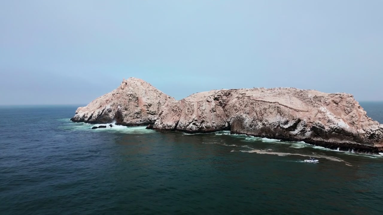 Riprese aeree orizzontali con drone che eseguono un'ampia orbita attorno a un'isola rocciosa al largo della costa di Lima, Perù. L'Oceano Pacifico cristallino circonda la formazione rocciosa naturale