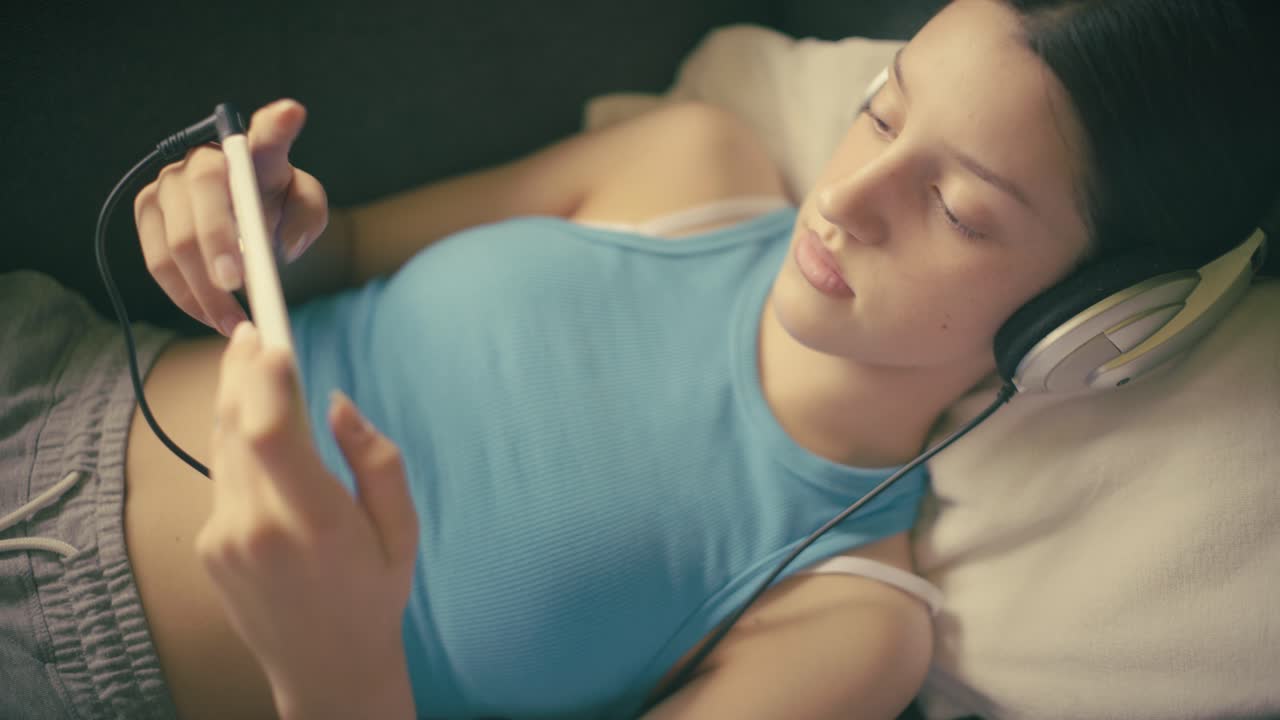 Teen girl relaxing at home, listening to music with headphones and using her smartphone. A calm, intimate moment of digital leisure in a cozy setting.