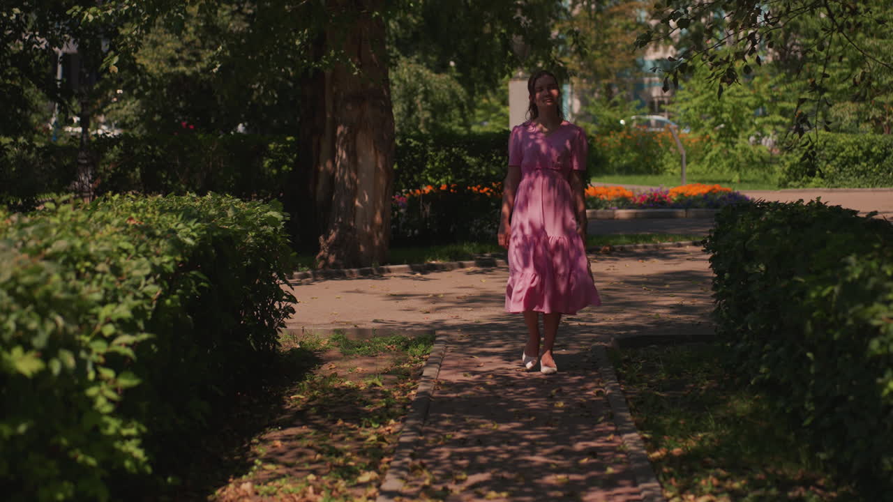 Donna che cammina lungo il sentiero soleggiato di un parco indossando un abito rosa, inquadrata da siepi curate e un albero alto, ombre chiazzate sul pavimento, foglie cadute sparse, passi lenti e misurati che trasmettono una sensazione romantica e pacifica