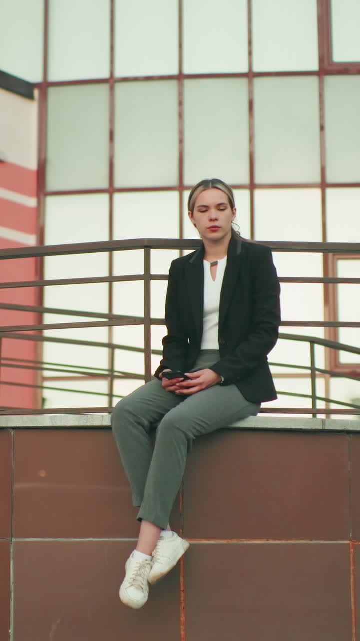 Lady in black blazer and white shirt seated on fence in urban area holding phone, waiting for someone, legs crossed at ankle, modern glass building in background