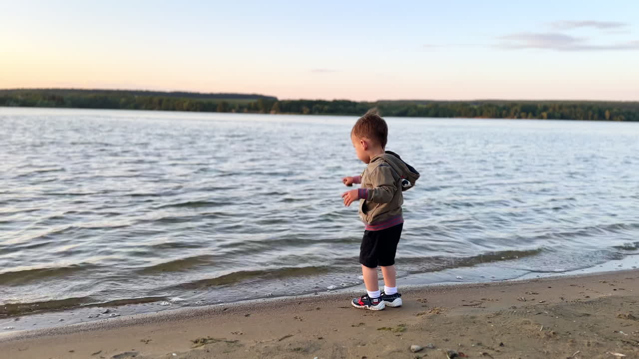 Adorable baby boy plays with on the shore at sunset. Kid takes some sand into hand and throws it into water.