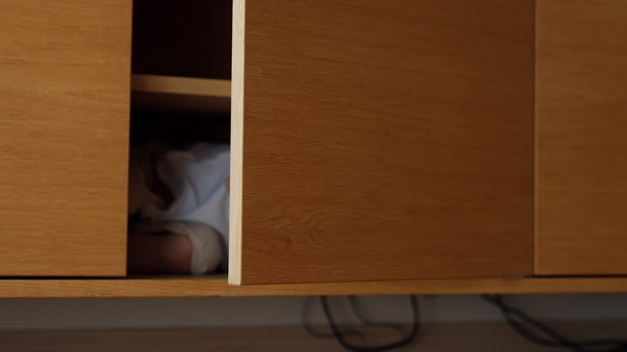 Joyful Caucasian baby boy in white t-shirt lies in the commode. Kid closes the doors hiding inside. Toddler playing at home
