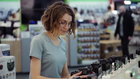 Mujer-Joven-Con-Gafas-Comprando-Una-Nueva-Cámara-Fotográfica-En-La-Tienda-De-Electrónica.-Tratando-De-Decidir-Cuál-Es-El-Mejor-Modelo.-Tiene-Dudas