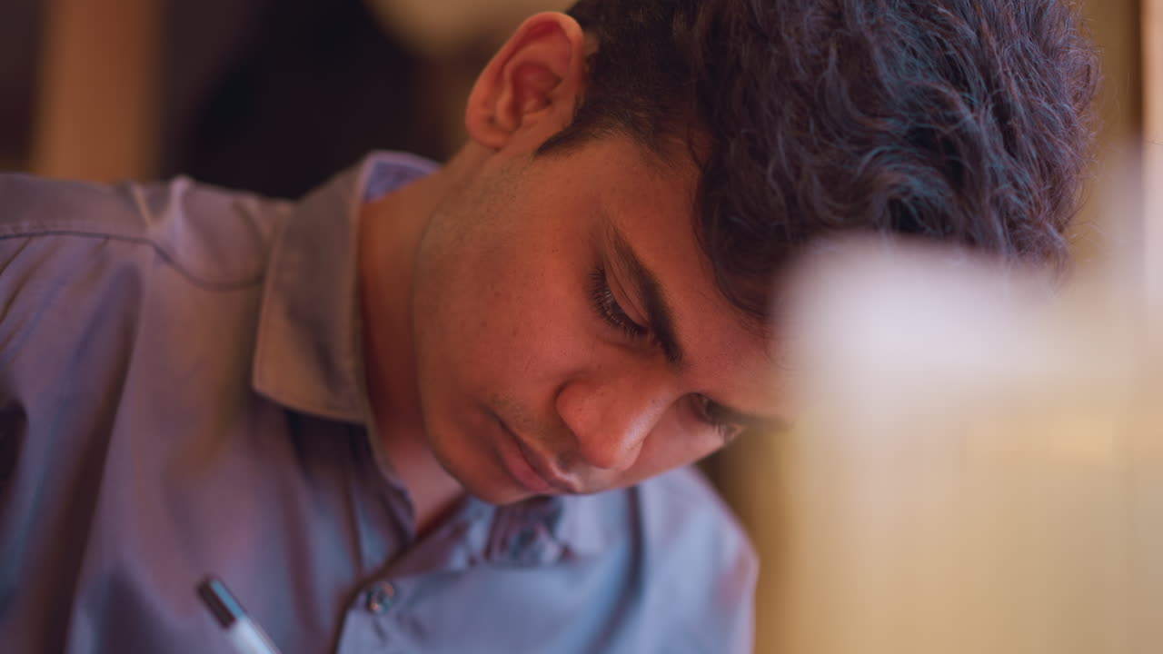 Young man with concentrated expression leans forward with downward gaze in warmly lit indoor setting, suggesting intense focus, artistic dedication, or thoughtful immersion in creative or academic activity