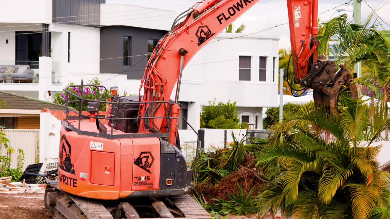 An orange hydraulic excavator removes a palm tree from a residential construction site in daylight, with steady camera and clear, natural lighting