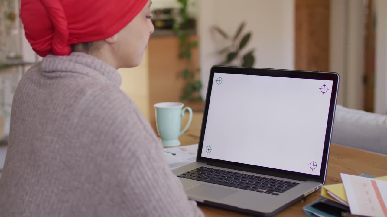 Video of biracial woman in hijab sitting at desk at home, working on laptop, copy space on screen