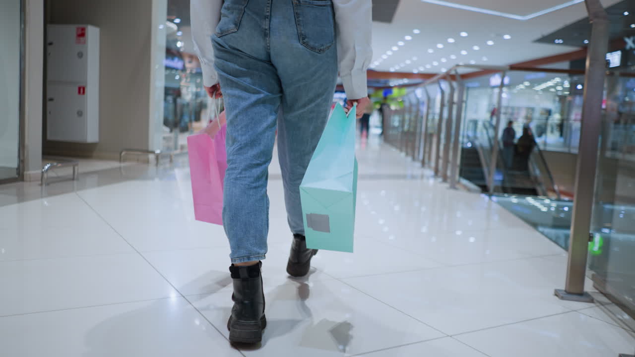 vista trasera de una mujer segura con gafas caminando a través de un centro comercial moderno, sosteniendo bolsas de compras de colores en ambas manos, ella camina a propósito en un brillante