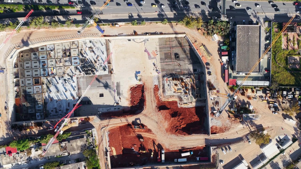 Majestuosa vista aérea cenital que revela una gran obra en construcción con grúas y movimientos de tierra, situada junto a una concurrida autopista urbana con tráfico constante bajo la brillante luz del sol.