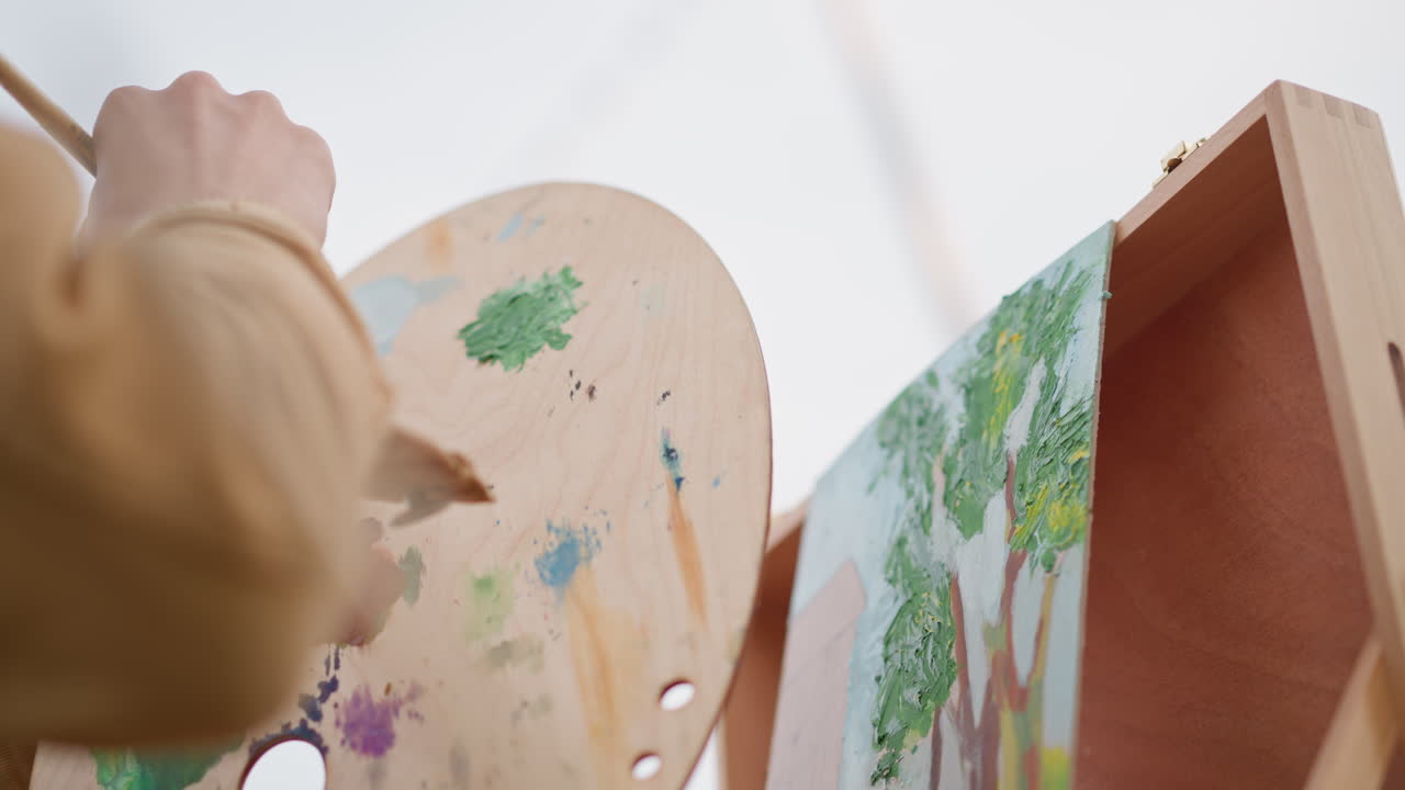 Dipinto con vista da sopra la spalla raffigurante lo studio di un albero, palette di legno costellata di verdi e viola mischiati, polso fermo che aggiunge consistenza ai tronchi e al fogliame, scatola con cavalletto portatile in uno studio luminoso, concentrato