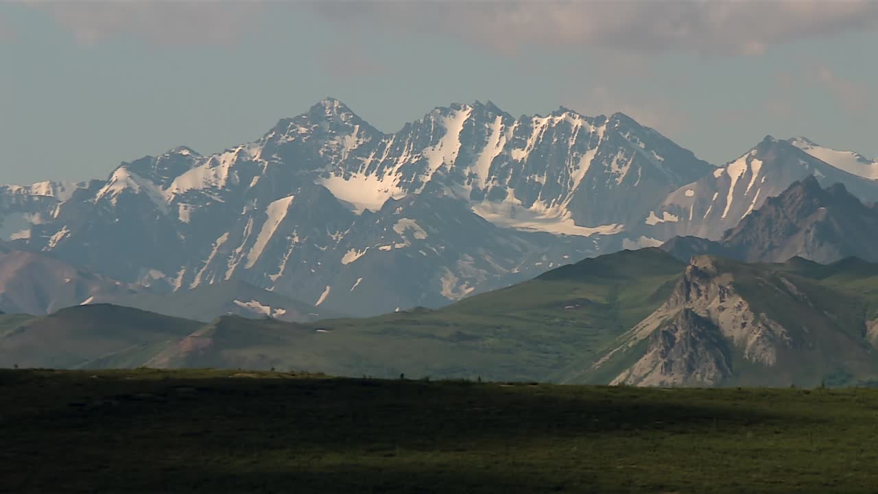 Snow covered peaks with rugged rocky ridges and green foothills in Denali National Park, Alaska. Static daytime view