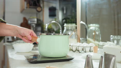 woman places mint green pot on electric stove in bright kitchen surrounded by eggs, glass jar of flour, colander, and utensils on counter, preparing to cook with modern and vintage kitchen elements