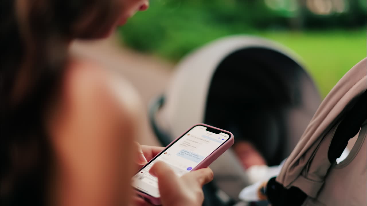 A woman with long curly hair texting on her phone while sitting on a park bench near a stroller, surrounded by greenery