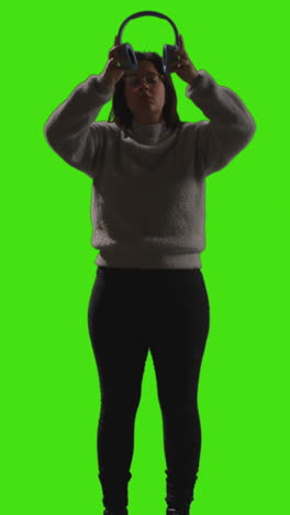 Vertikale-Videoaufnahme-Einer-Frau-Mit-Kabellosen-Kopfhörern,-Die-Musik-Vom-Mobiltelefon-Streamt,-Vor-Einem-Greenscreen-Mit-Schwacher-Beleuchtung