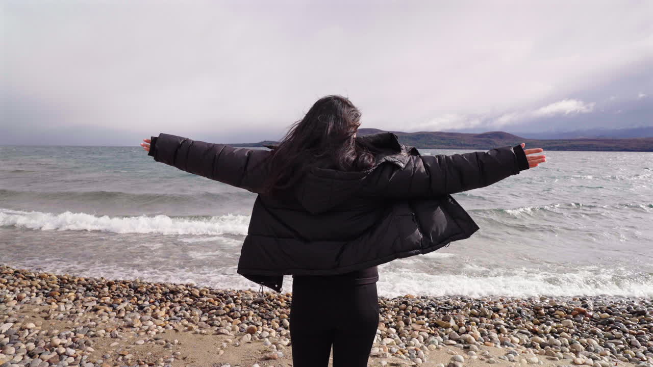Woman standing with arms wide open facing and thinking of her personal freedom in front of lake waves on a cloudy and windy day.