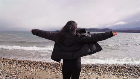 woman standing with arms wide open facing and thinking of her personal freedom in front of lake waves on a cloudy and windy day.