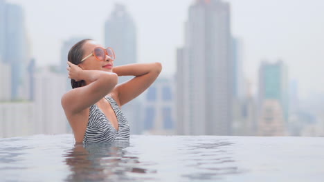 Una-Joven-Asiática-Apasionada-Toca-Su-Cabello-Mojado-Estando-Dentro-Del-Agua-De-La-Piscina-Infinita-En-La-Azotea-De-Un-Hotel-De-Lujo-Con-Rascacielos-De-La-Ciudad-En-El-Fondo