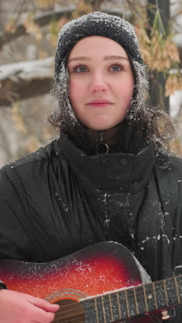 mujer con capucha negra tocando guitarra acústica al aire libre en un parque nevado, rodeada de árboles desnudos helados y nieve que cae suavemente, capturando el estado de ánimo sereno del invierno y la armonía entre la música