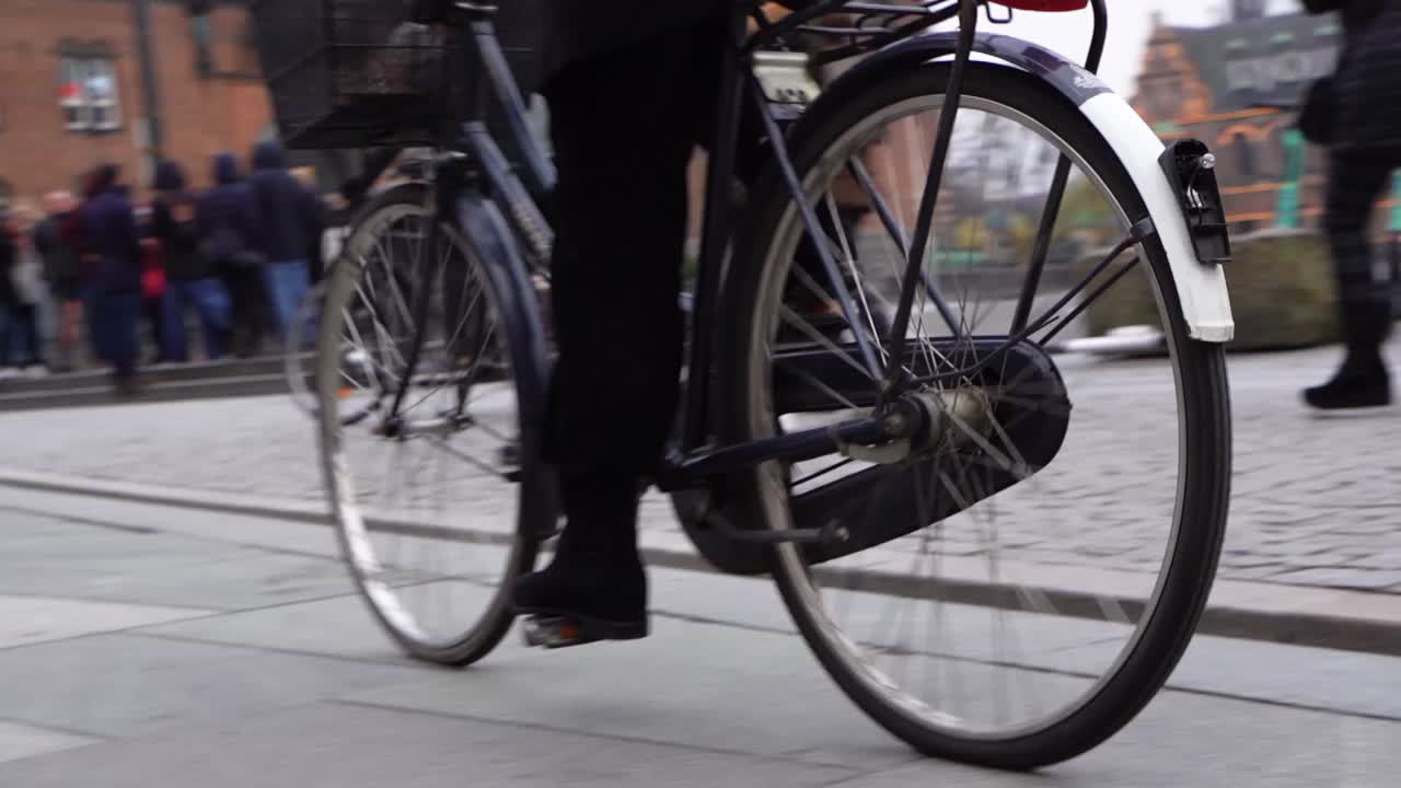 Person peddles bicycle on city sidewalk toward and past camera, close up slow motion low view, Copenhagen, Denmark