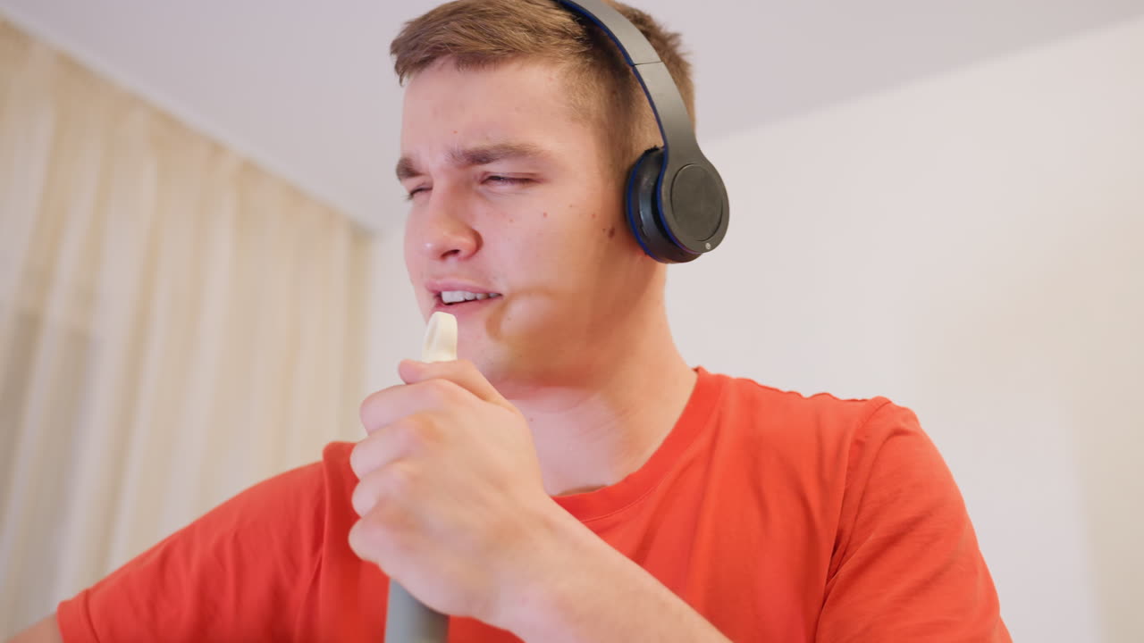 Young man in orange shirt wearing headphones sings with mop handle like microphone, eyes closed and arm raised, enjoying rhythm with expressive playful energy in bright home environment