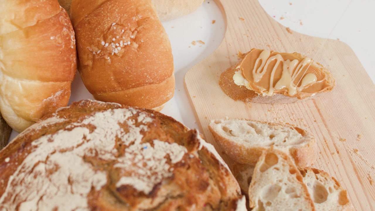 Creamy peanut butter drizzles onto crusty bread slices amidst assorted artisan bakery loaves.