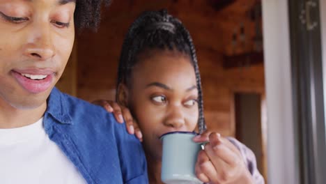 Feliz-Pareja-Afroamericana-Mirando-Por-La-Ventana-Y-Tomando-Café-En-Una-Cabaña-De-Madera,-Cámara-Lenta