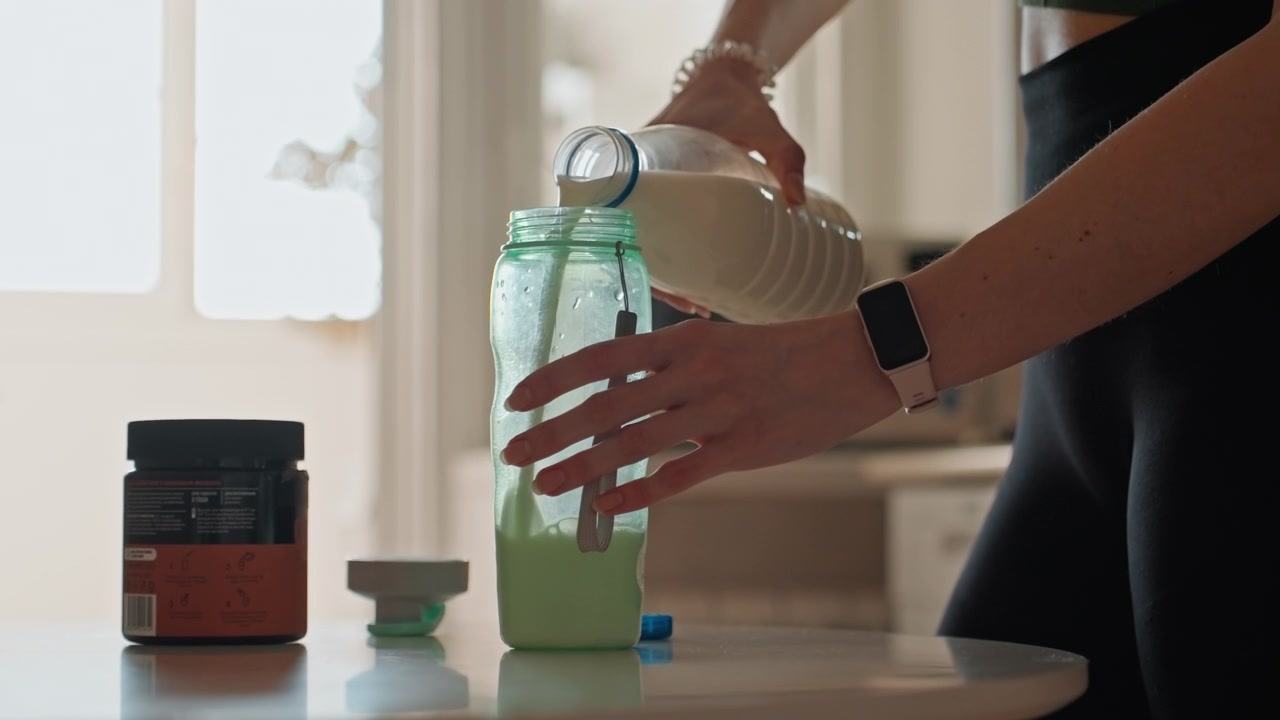 Home kitchen fitness woman pouring milk into green shaker jar beside supplement tubs, soft natural light, calm morning routine, closeup hands with watch, black leggings, mix and hydrate before workout