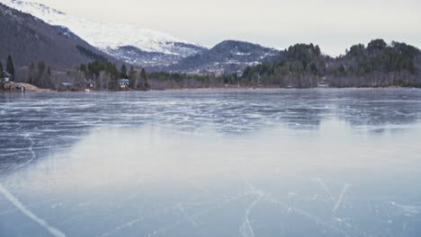 Camera-flying-over-a-frozen-lake,-revealing-beautiful-scenery-of-snow-covered-mountains-and-forest