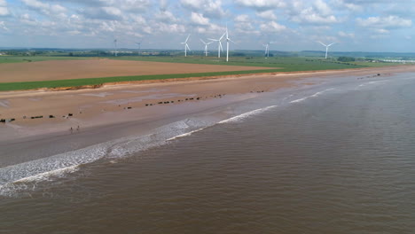 Establishing-Aerial-Drone-Shot-Over-Beach-with-Calm-Waves-and-Fields-with-Wind-Turbines-on-UK-East-Coast