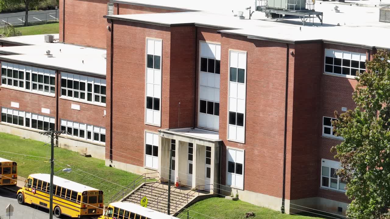 Pick up Line of yellow school buses in front of red brick school on american. Sunny day in fall season. Aerial zoom shot