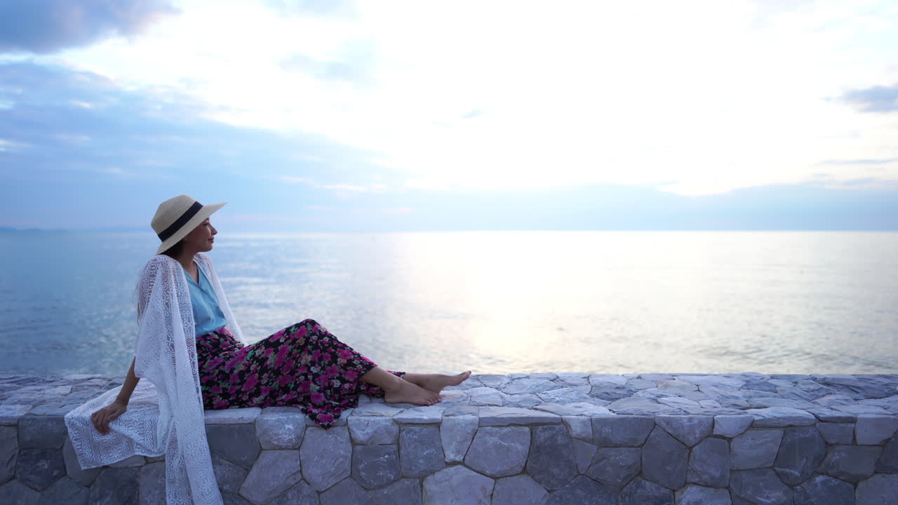 세련된 아시아 여성이 바다 성벽에 앉아 수평선을 바라보며 평화롭게 즐기고 있으며, 복사 공간 샷으로 정적입니다.