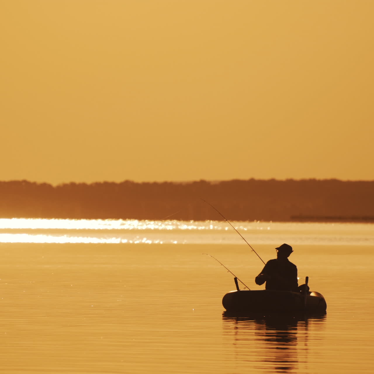 Silhouette of a man in hat fishing from the boat on the evening river background. Beautiful sunset and a fisherman catching fish from the inflatable boat in the open water.
