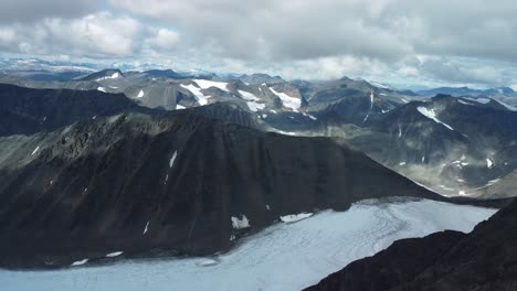 aerial drone footage flying over tall black and dark colored kebnekaise mountain tops that have white snow patches between the steep slopes and cliffs. sun is shining and creating sun patches, shadows