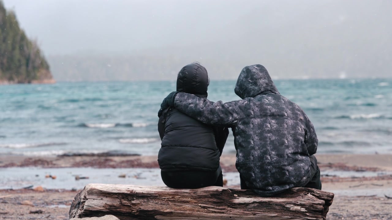 Two individuals sit closely on a log by the sea, one offering comfort with an arm around the other. The scene conveys a sense of companionship and support.