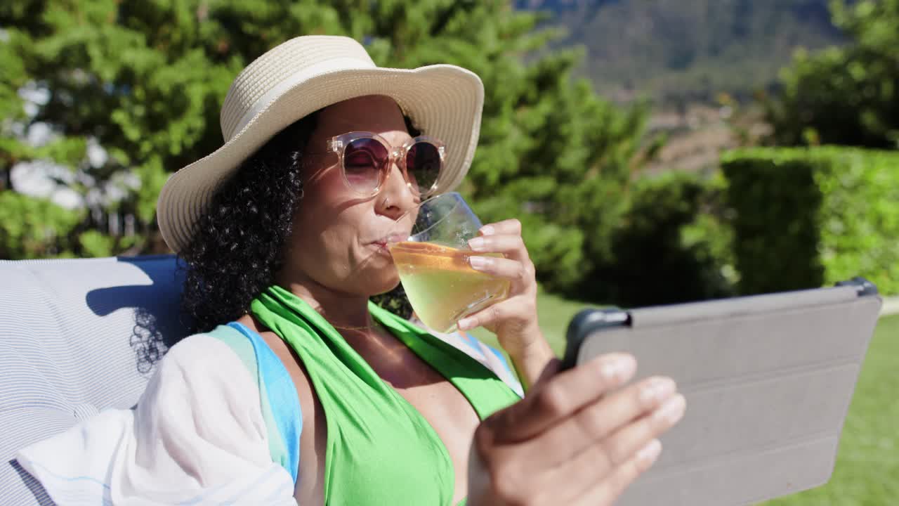 African American woman in sunhat reclining on lawn chair, tapping tablet, sipping glass with orange
