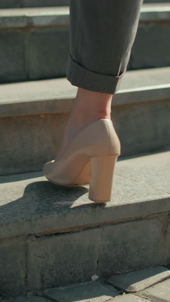 Lower leg view of woman walking in high heels on interlocked pavement ascending concrete steps, sunlight casting sharp shadow, focus on movement, footwear, business attire, and urban setting
