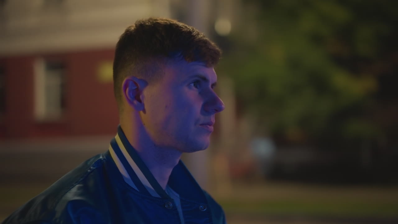 White young man drinking at night by park, close profile shots with soft bokeh and neon highlights, wearing varsity jacket and jeans, looking into distance in thoughtful pose, scenes suggest student
