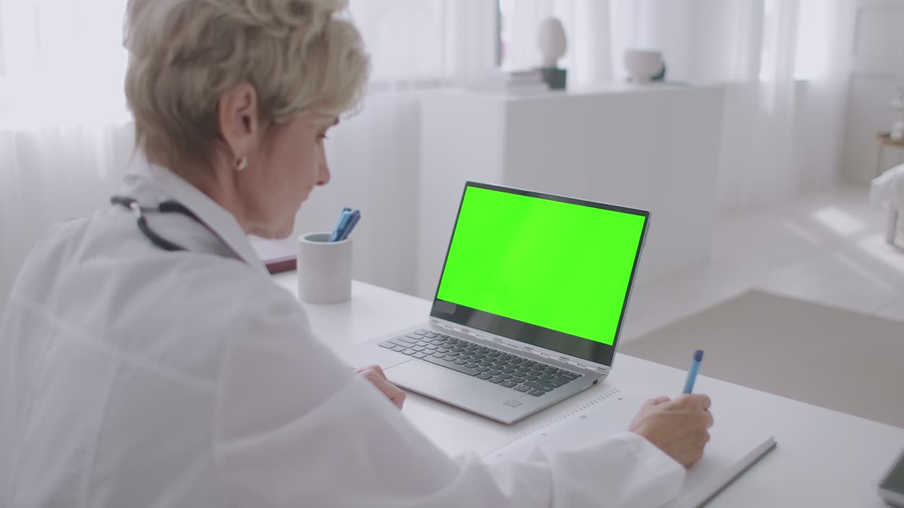 mujer médico está viendo webinar, mirando la pantalla verde de la computadora portátil para la tecnología de clave de croma, escribiendo notas en el cuaderno, capacitación avanzada
