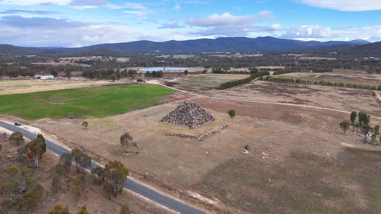 Drone glides over a dry rural landscape, circling a large man-made stone pyramid with distant hills and scattered trees under bright daylight