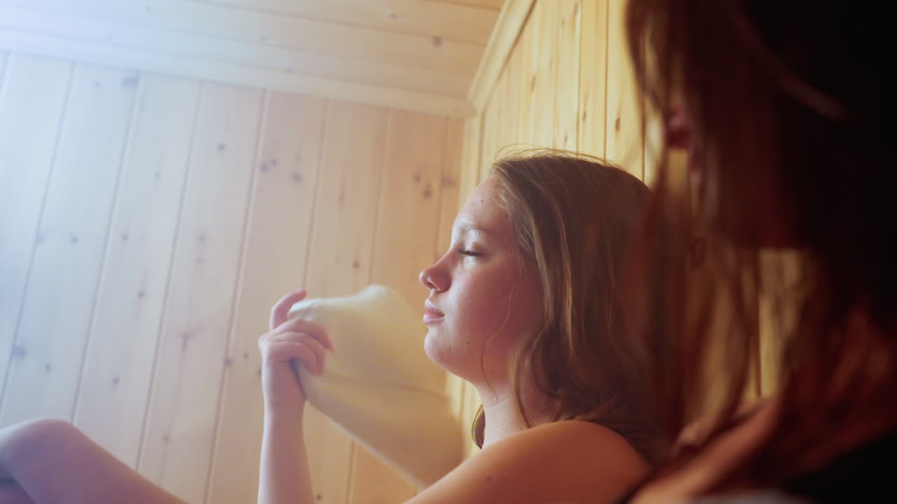 close up of woman seated next to sister adjusting hair and fanning with felt hat in steamy sauna cabin, warm wooden walls, relaxed mood and subtle steam haze enveloping scene, calm expressions