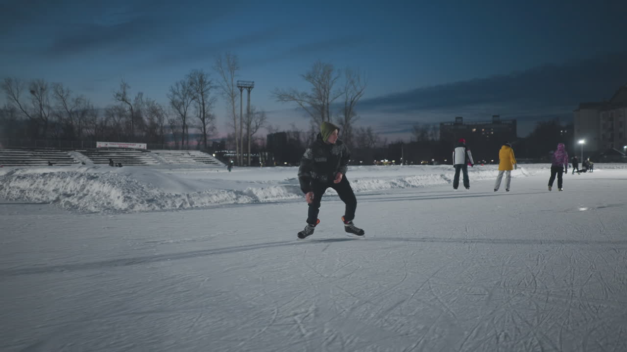 professional skater gliding skillfully across outdoor ice rink at night wearing patterned winter jacket surrounded by other skaters with urban buildings and bright stadium lights in background