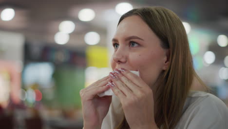 young woman sitting alone at table in mall dining area cleaning her mouth with napkin, enjoying relaxed moment, background features colorful lights