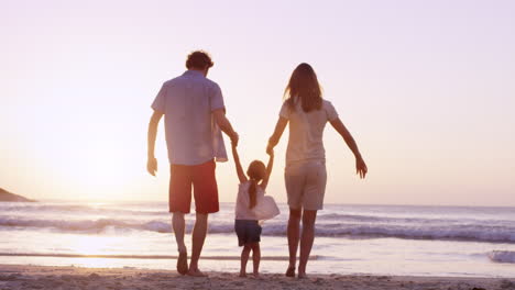 Glückliche-Familie-Am-Strand,-Die-Händchen-Hält-Und-Ein-Kleines-Mädchen-Bei-Sonnenuntergang-Im-Urlaub-Herumschwingt