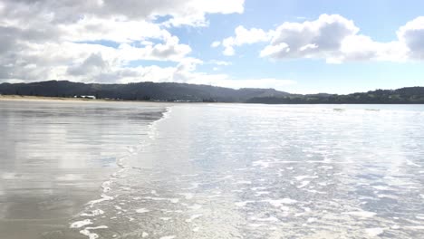 Shallow-wave-flowing-in-diagonally-onto-a-beach-against-a-mountain-background-during-the-daytime