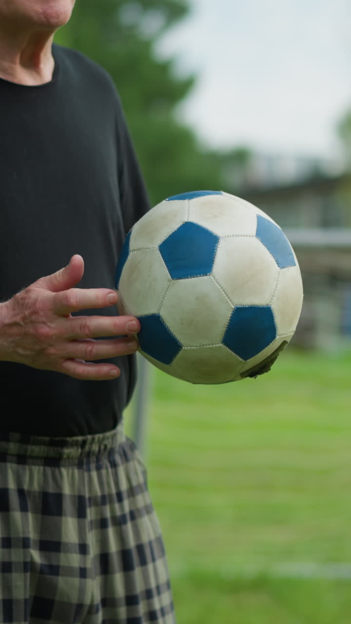 An elderly man rolls a soccer ball back and forth in his hand with focused intent, in the blurred background, a goalpost and a building can be seen