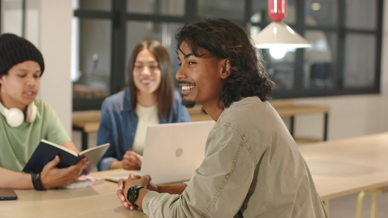 Homme adulte entamant une discussion pour une étude à une table avec un ordinateur portable, gesticulant, se tournant vers la caméra, souriant