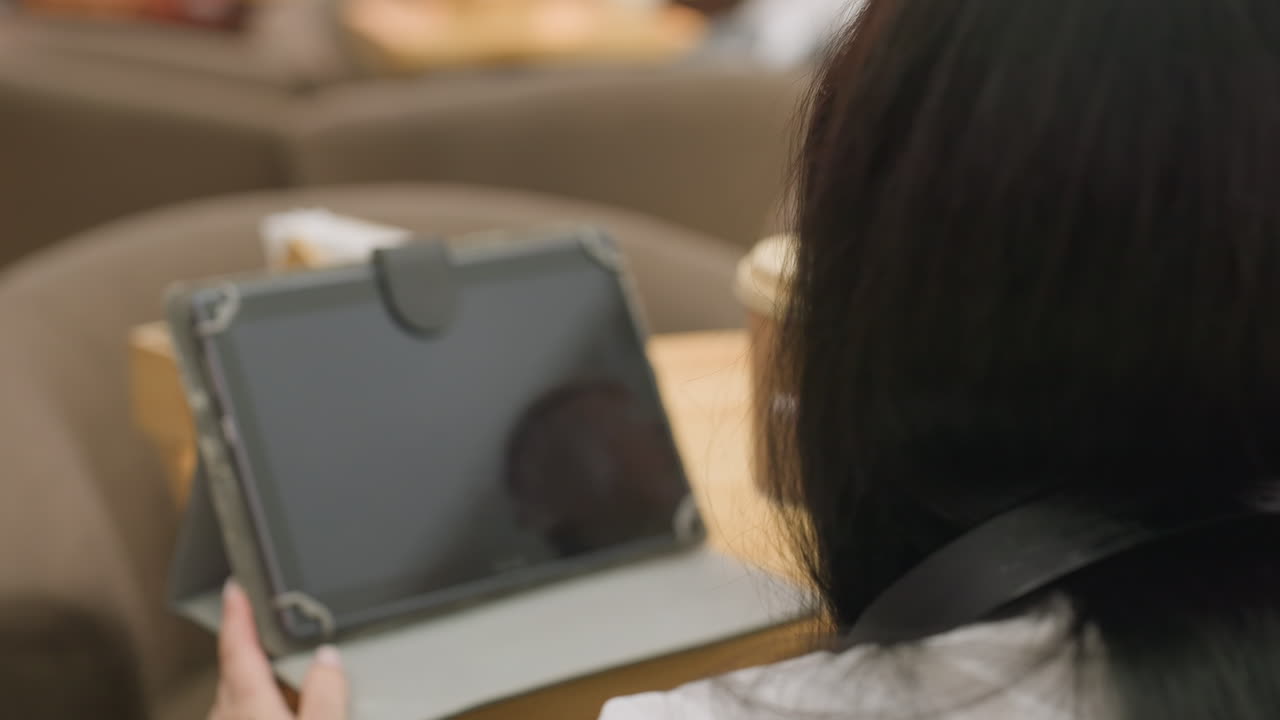 close up of woman using tablet with one hand while other holds disposable juice cup seated at cozy indoor cafe setting with warm lighting and relaxed atmosphere during work session