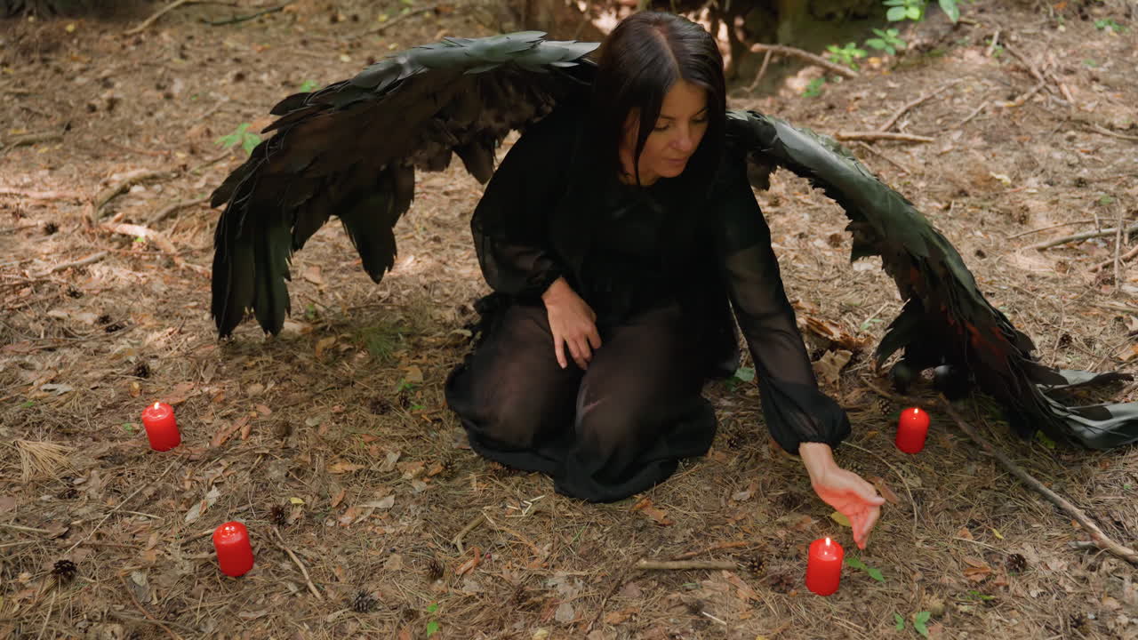 Sorceress with black wings kneels on forest ground surrounded by red candles, touching flame from left side before beginning prayer to forest spirit, evoking mystical and spiritual atmosphere